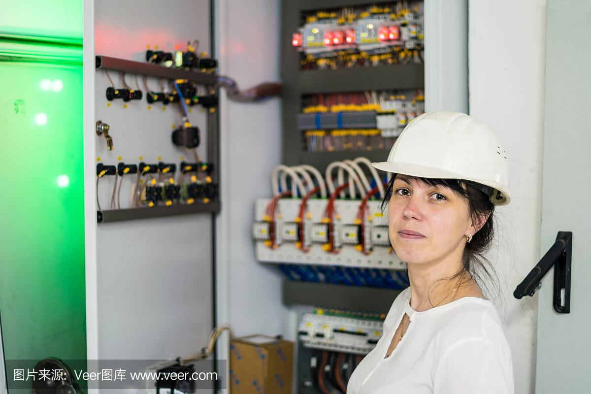 一名戴着白色头盔的电工妇女的肖像,旁边是高压电气设备。一个戴着工程头盔的女孩站在保险丝盒的背景上。